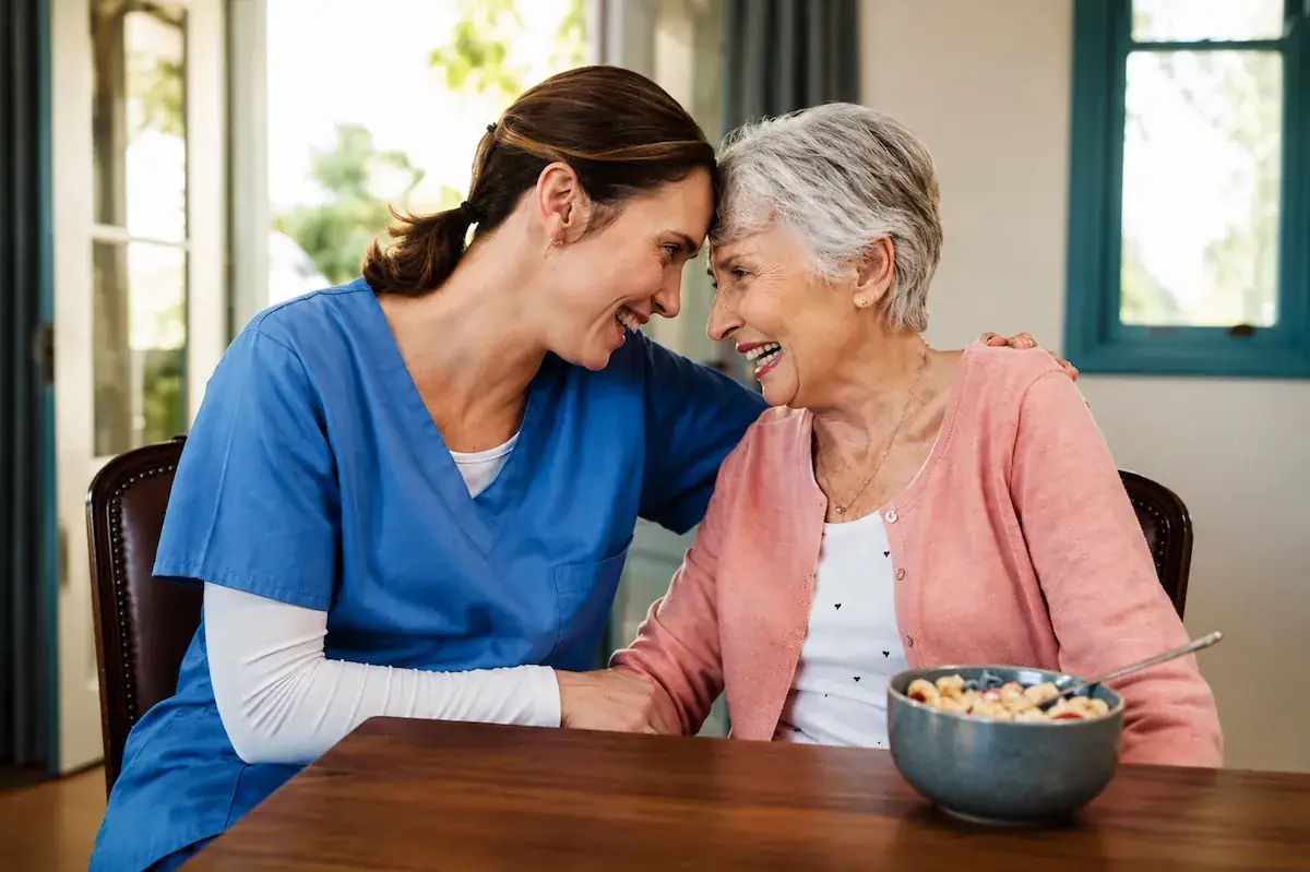 A caregiver and a senior woman sharing a laugh at the kitchen table